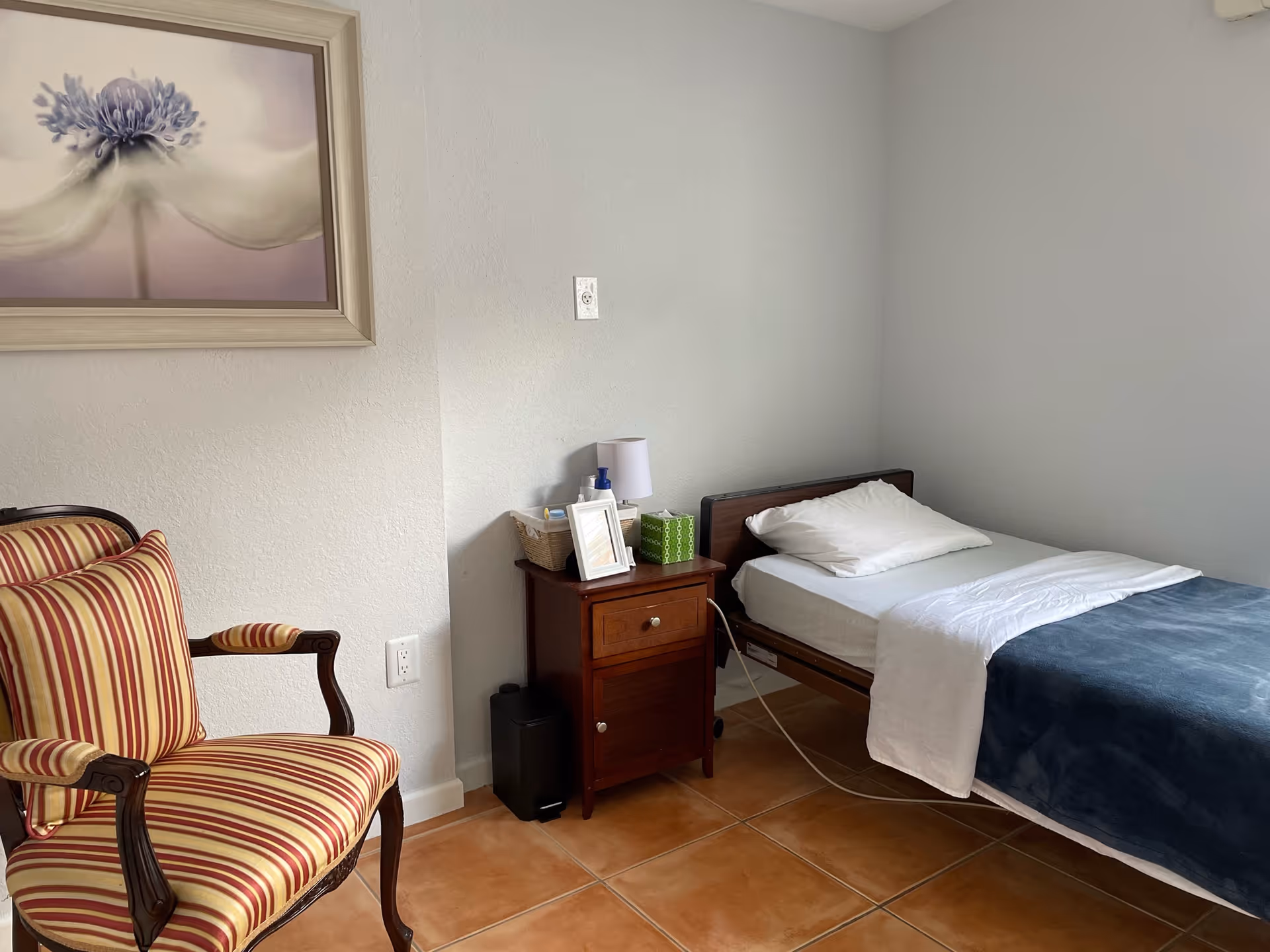 A simple bedroom with a single bed covered with white and dark blue bedding, a wooden nightstand with a lamp, tissue box, and framed photo, a striped armchair with red and beige colors, and a large framed flower painting on the wall.