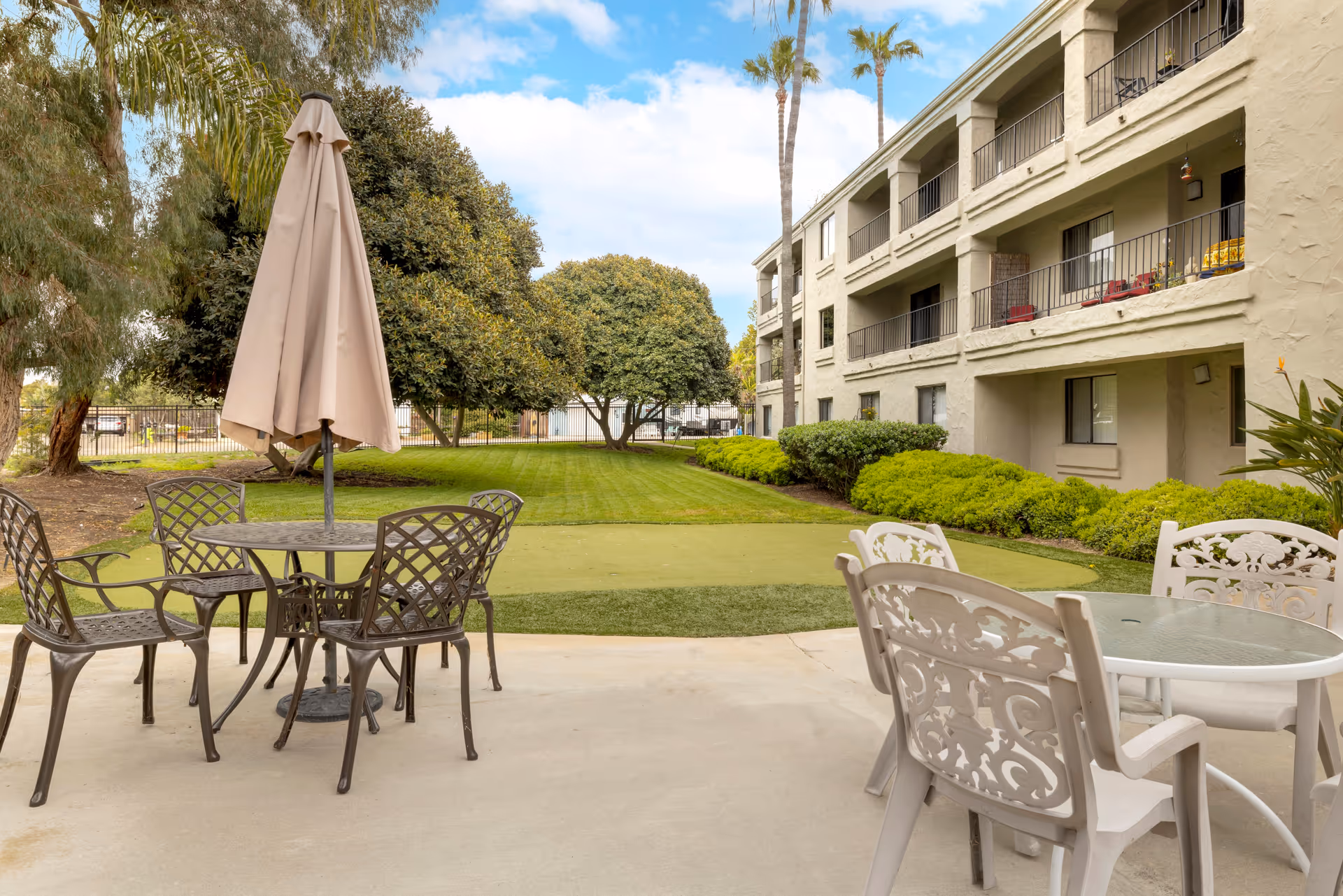 Outdoor patio with metal tables and chairs, umbrella, and a multi-story beige building.