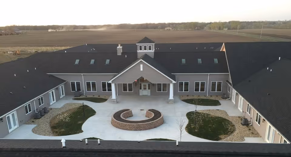 Aerial view of the Villas of Holly Brook - Morton senior living facility showing a large U-shaped building with a central entrance covered by a gabled roof. The courtyard features a circular brick seating area surrounded by concrete walkways and landscaped patches of grass and rocks. The building has multiple windows and skylights on the roof, with open fields visible in the background.