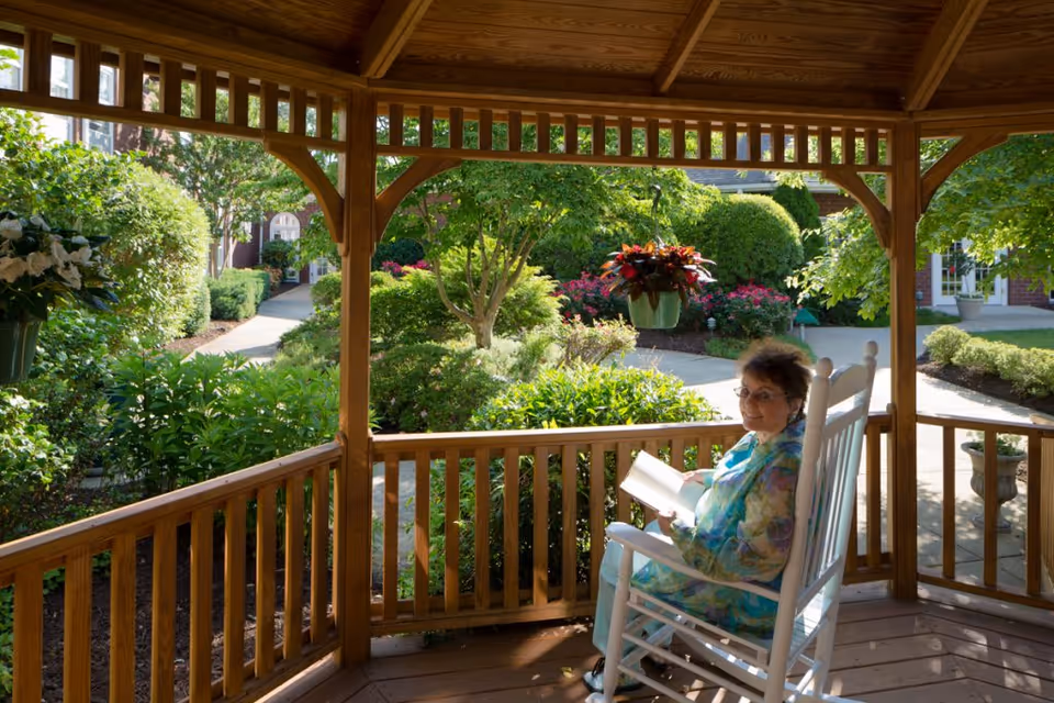 An elderly woman sitting in a white rocking chair on a wooden gazebo porch, reading a book. The gazebo overlooks a lush garden with green bushes, trees, and colorful flowers, with a paved walkway and building visible in the background.