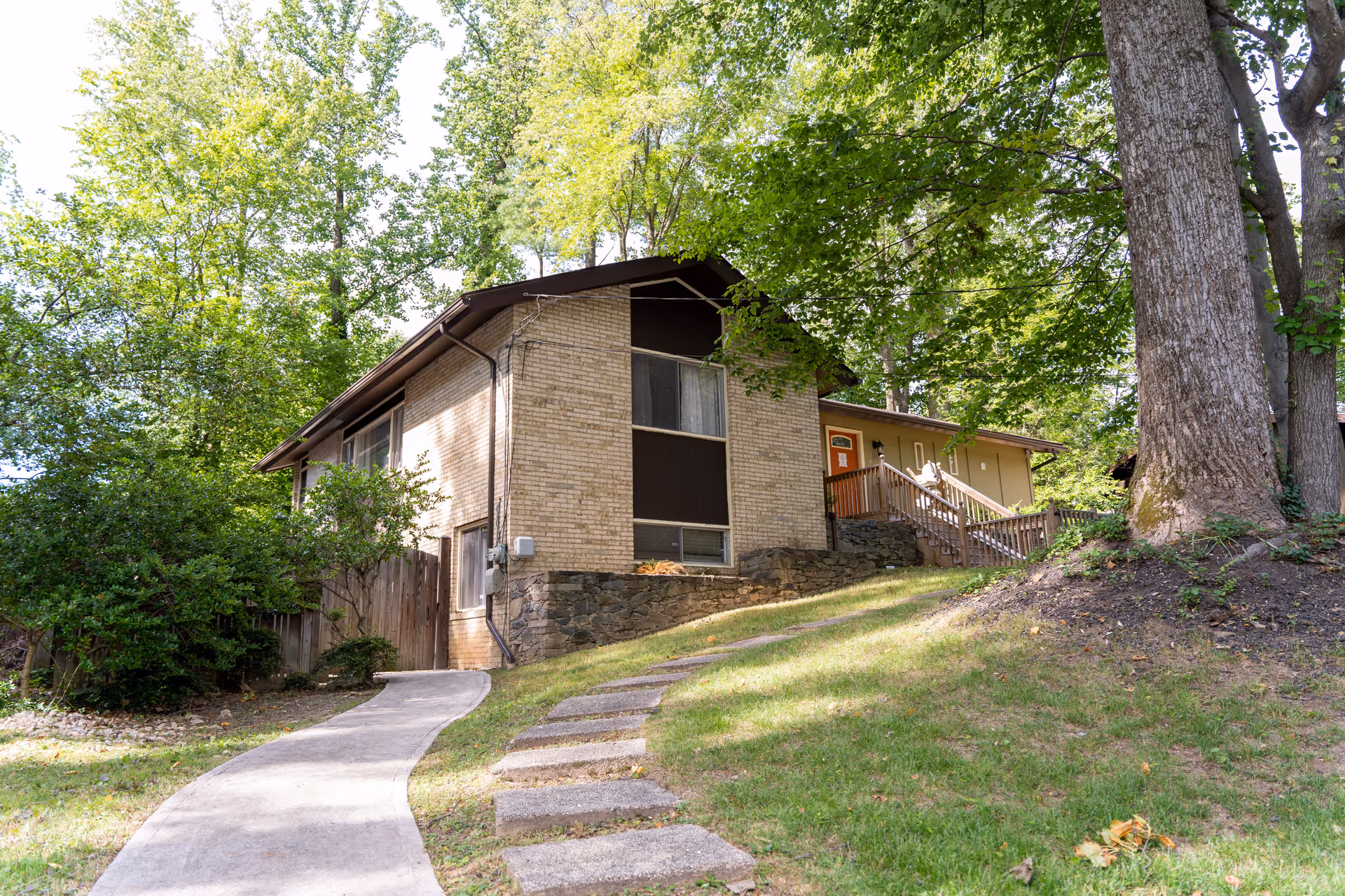 Brick residential building with a curved concrete walkway and steps leading to a porch, surrounded by trees on a sloped lawn.