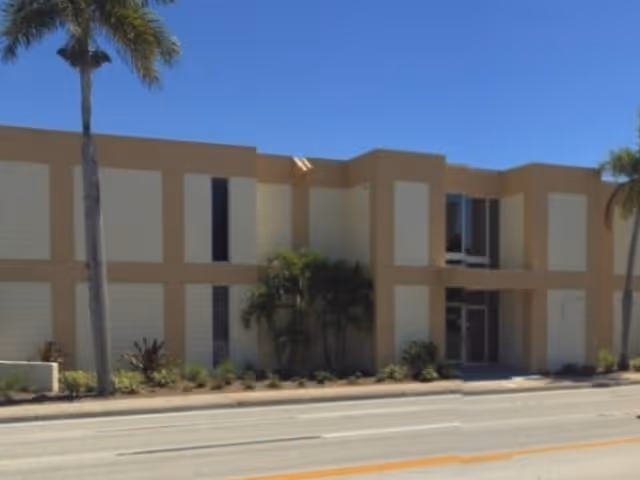 Beige two-story building with palm trees and a street in front