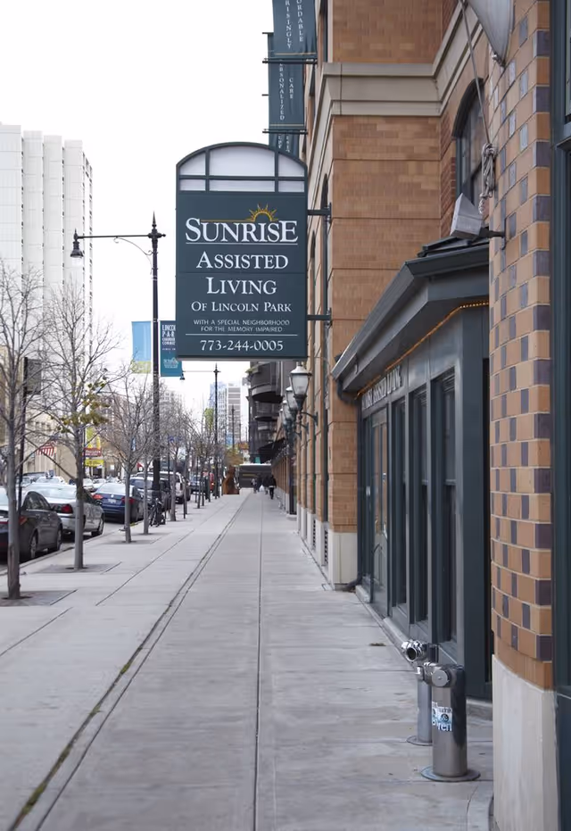 Sidewalk view along a city street with parked cars and leafless trees. A large hanging sign on a brick building reads 'Sunrise Assisted Living of Lincoln Park' with a phone number and additional text about memory care. The building has large windows and street lamps along the sidewalk.