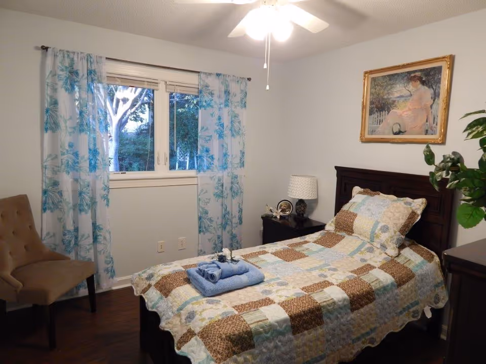 Bedroom in Bright Horizon Senior Care featuring a single bed, patchwork quilt, and blue curtains.