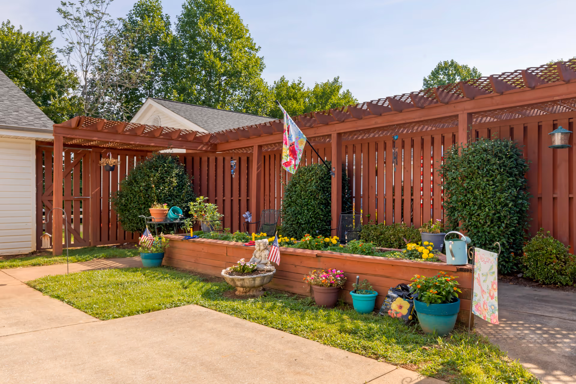 Outdoor garden area with a raised wooden planter box filled with flowers and plants, surrounded by potted plants and garden decorations. There is a wooden pergola and fence in the background, with green trees visible behind the fence.