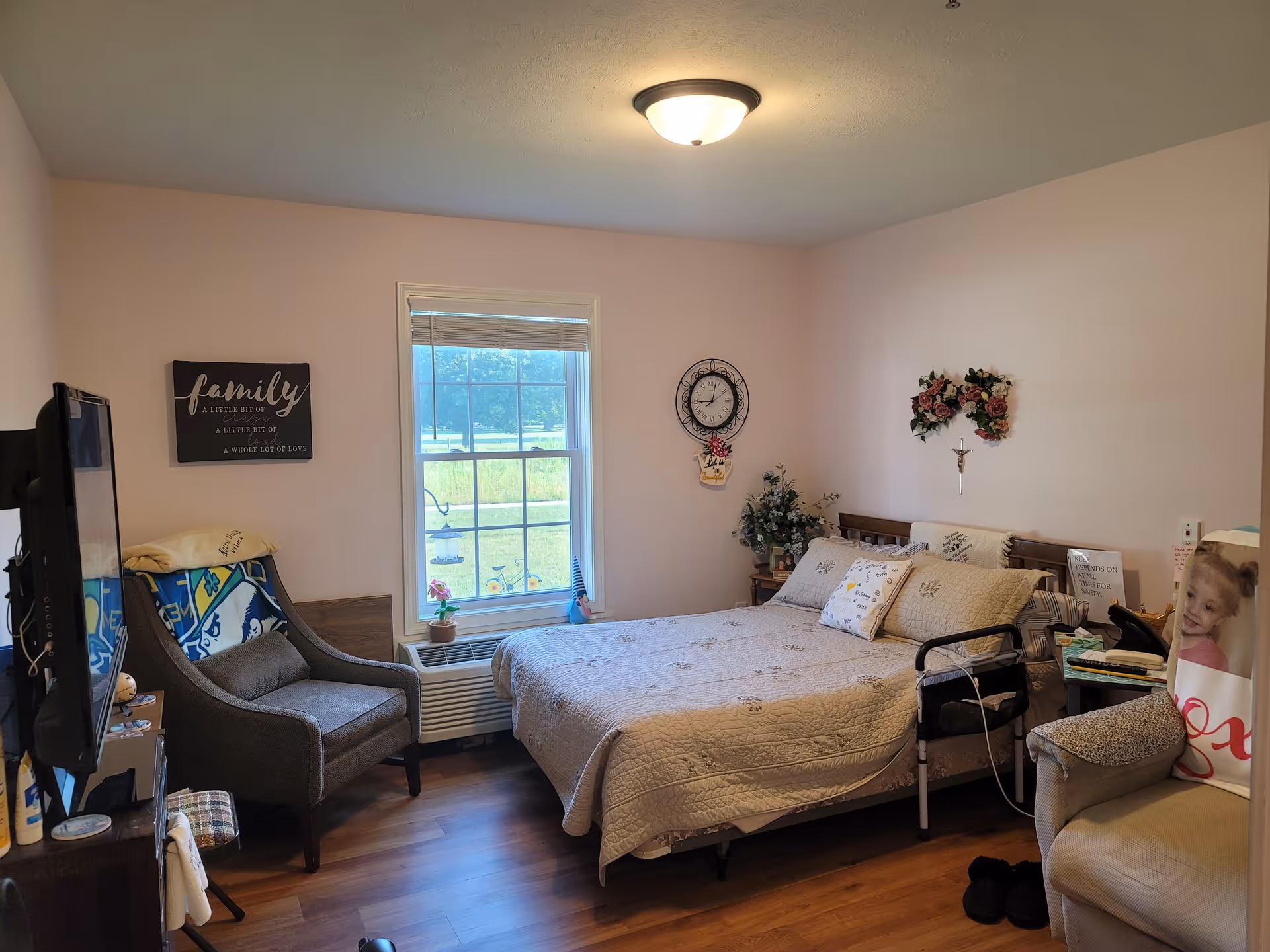 A cozy bedroom with a bed covered in a beige quilt and multiple pillows. There is a window with a view of greenery outside, a gray armchair with a blanket draped over it, a wall clock, floral wall decorations, and a small side table with a walker next to the bed. The room has wooden flooring and soft lighting from a ceiling fixture.