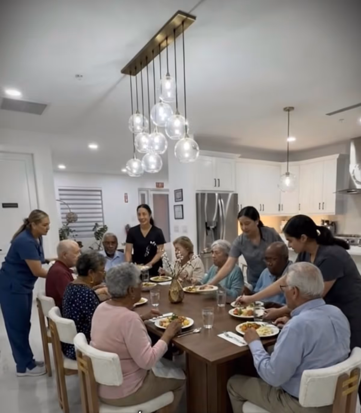 Group of elderly residents and caregivers dine at a long wooden table in a bright kitchen.