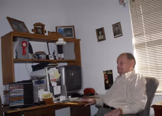Senior resident sits at a cluttered desk in a small home office within the facility.