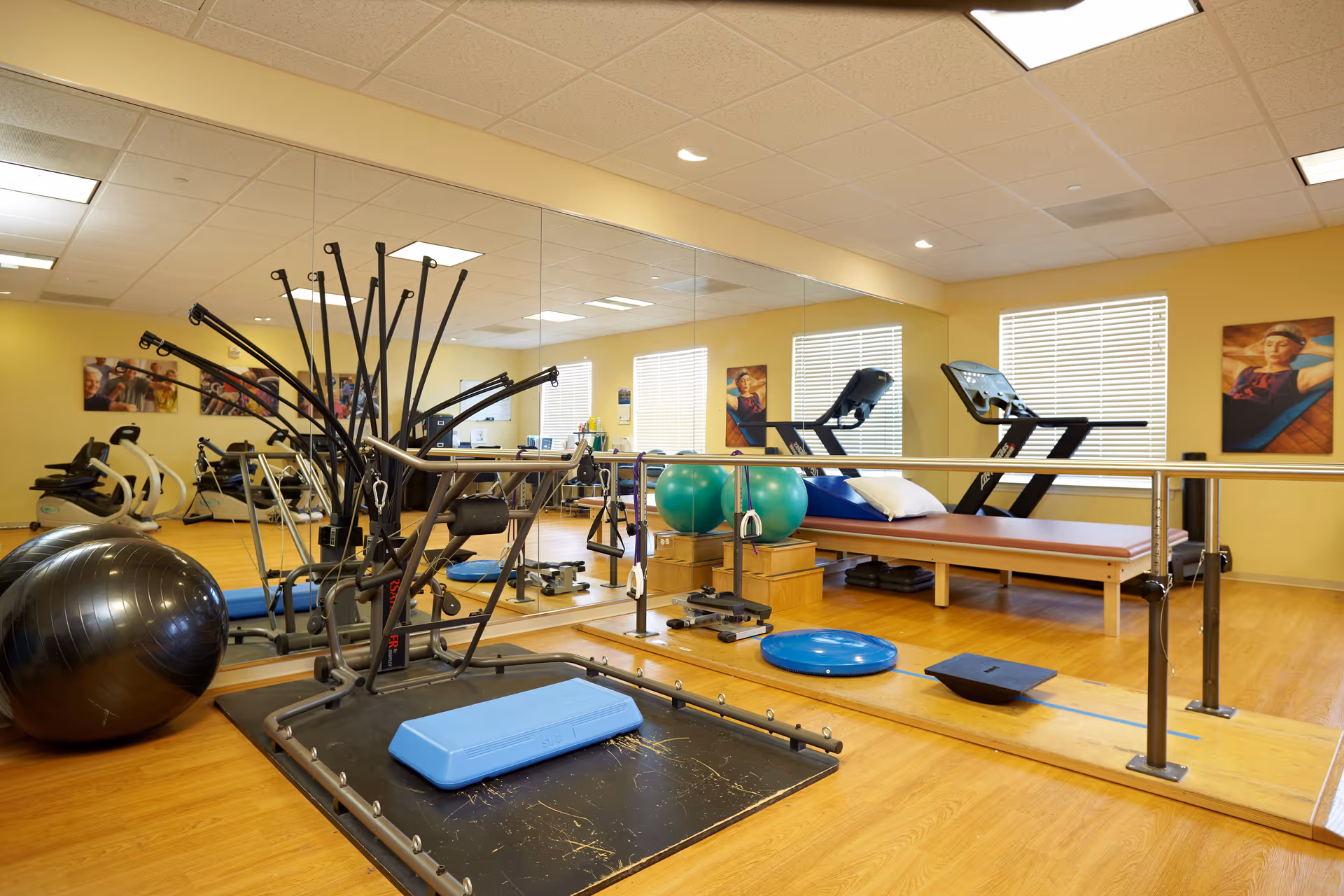 A senior living facility exercise room with various fitness equipment including a large exercise ball, resistance training machine, balance boards, and a treadmill. The room has wooden flooring, yellow walls, large mirrors, and windows with blinds. There are also framed pictures on the walls.