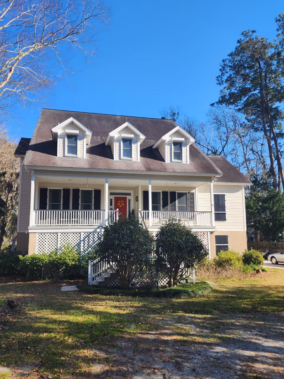 A two-story house with a wraparound porch, white railings, shrubs, and a clear blue sky.