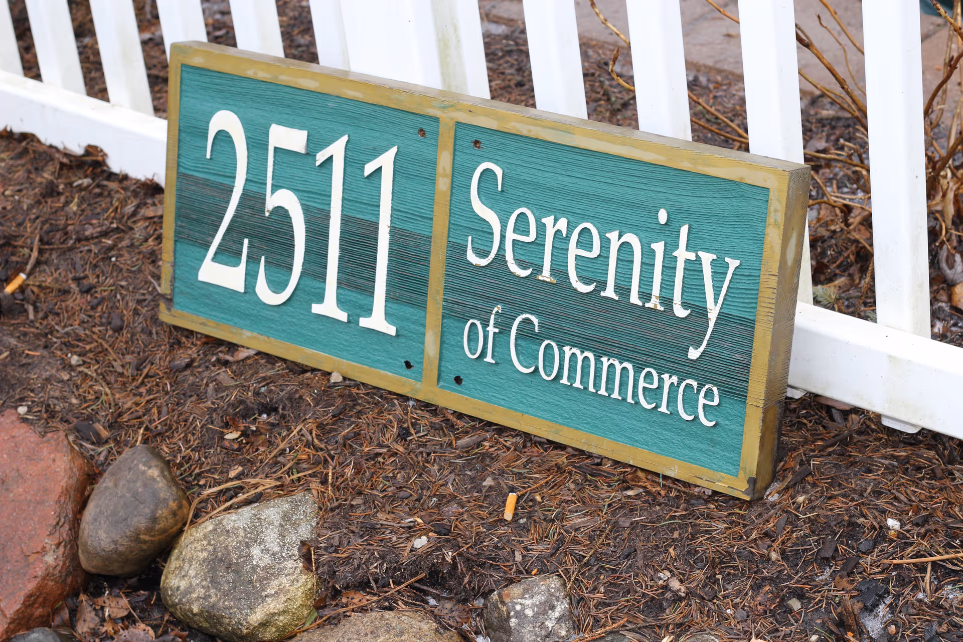 Green wooden sign reading 2511 Serenity of Commerce rests on mulch beside a white fence.