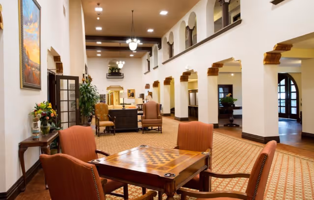 Spacious common room in a senior living facility with a game table and upholstered chairs, high ceilings, arched walkways, and seating areas.