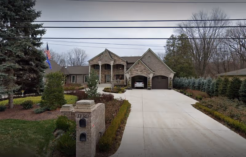 Front exterior view of a large brick house with a wide driveway leading to a two-car garage. The house has a covered porch with columns and is surrounded by landscaped bushes, trees, and a lawn. An American flag is visible on the left side near the driveway.
