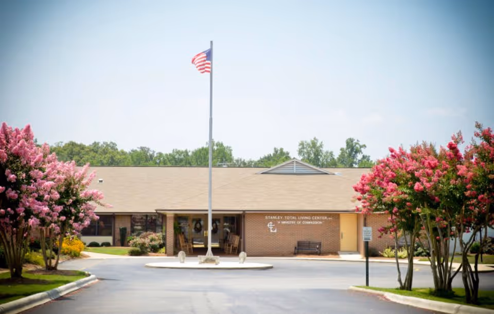 Front exterior view of Stanley Total Living Center building with an American flag on a flagpole in the center, surrounded by pink flowering trees and a paved driveway.
