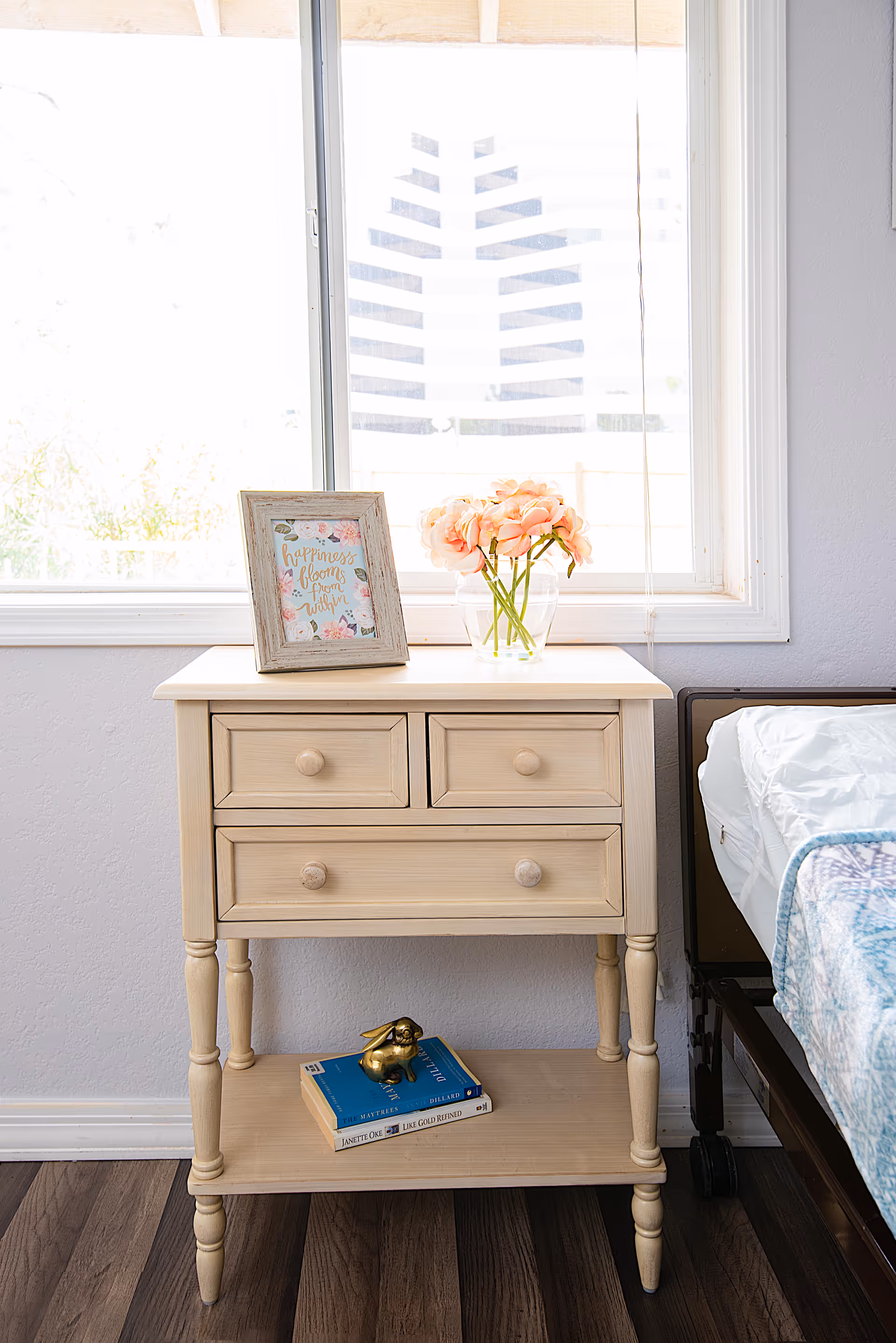 Bright bedroom with a cream nightstand, pink flowers, and a bed by the window
