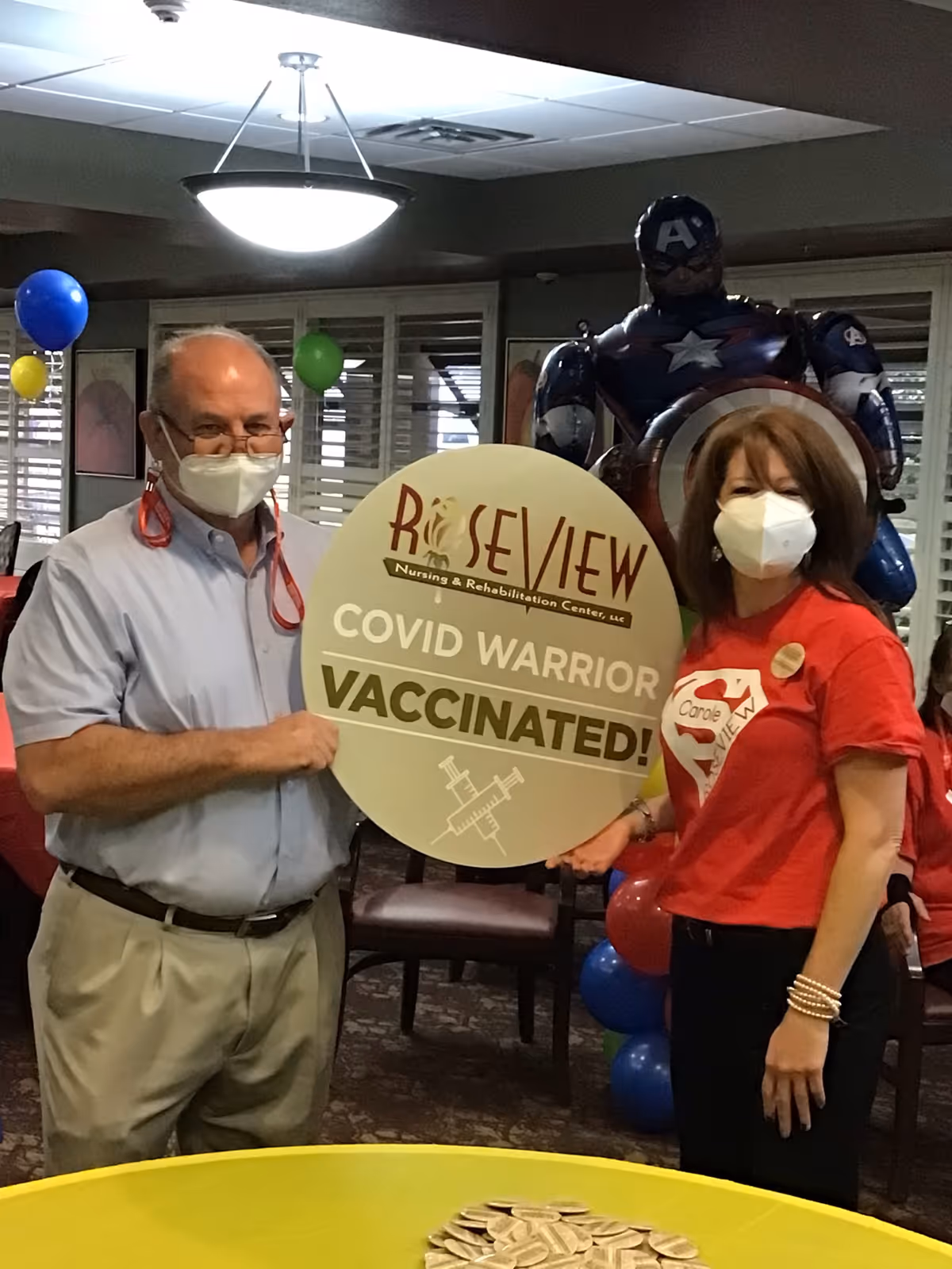 Two people wearing face masks stand indoors holding a large circular sign that reads 'Roseview Nursing & Rehabilitation Center, LLC COVID WARRIOR VACCINATED!' Behind them is a Captain America balloon figure and colorful balloons in the background.