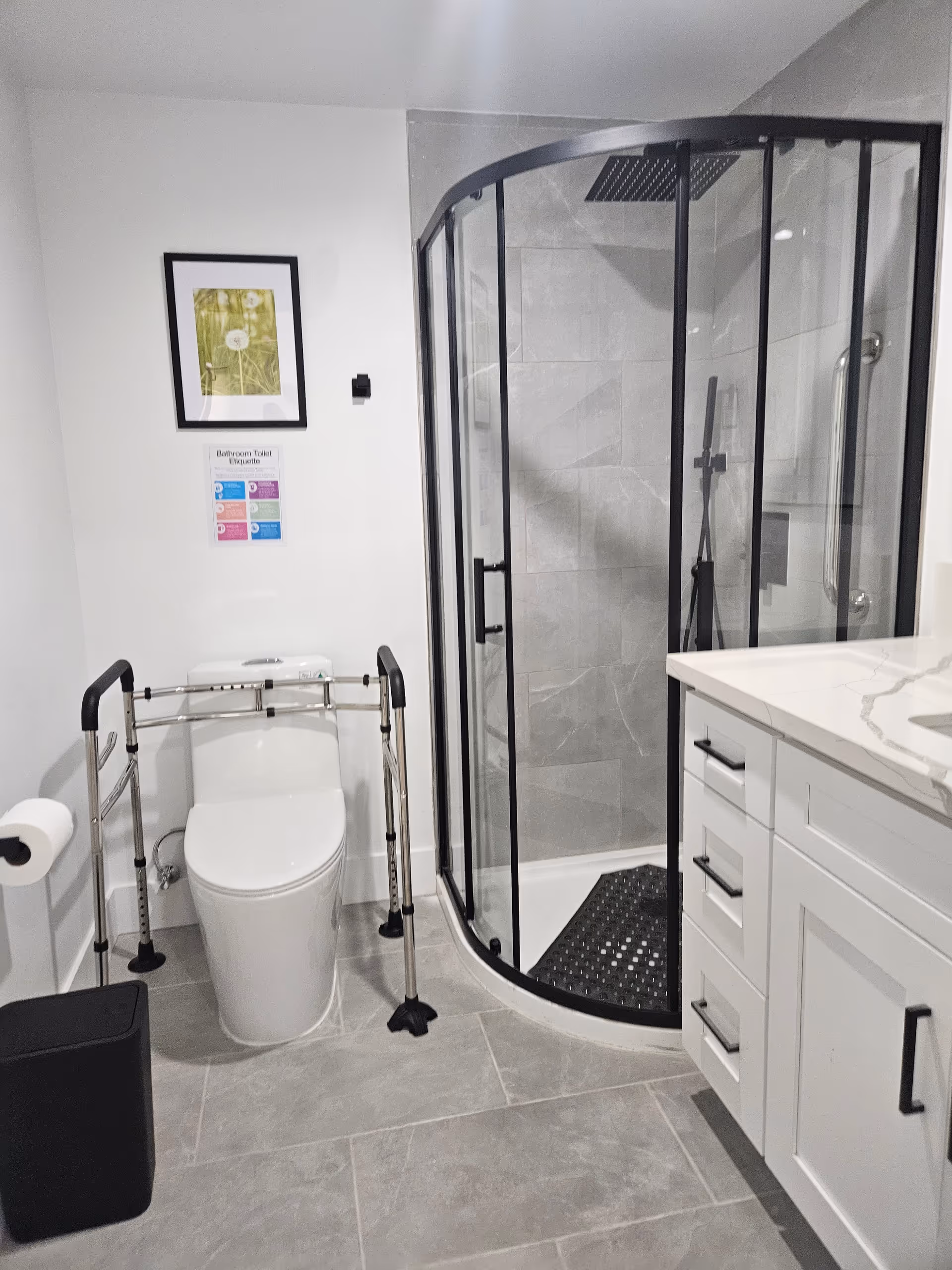 Modern bathroom with curved glass shower, toilet with grab bars, and white vanity.