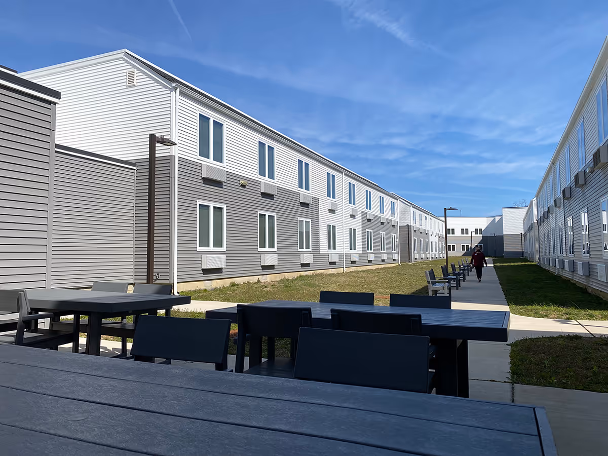 Outdoor courtyard area between two long, two-story buildings with multiple windows. There are several black tables and chairs in the foreground and a paved walkway with benches along the right side. A person is walking down the path under a clear blue sky.