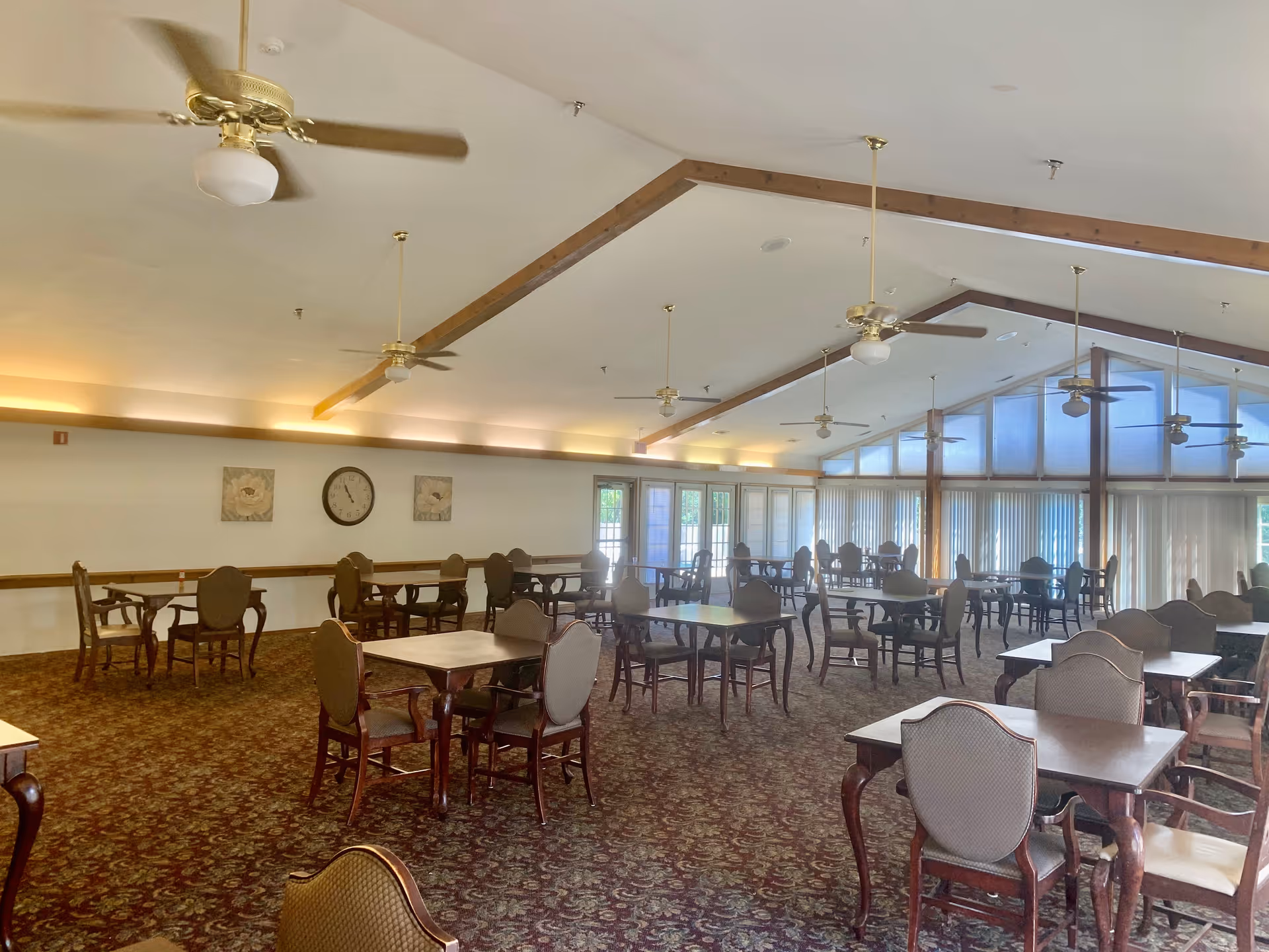A spacious dining room with multiple wooden tables and cushioned chairs arranged neatly. The room has a patterned carpet, ceiling fans, and large windows with vertical blinds allowing natural light to enter. The ceiling features wooden beams and soft lighting along the walls.