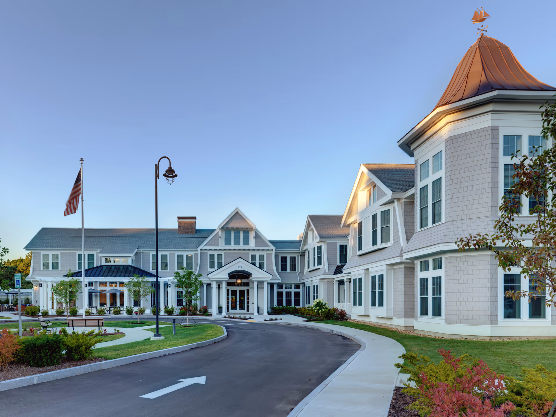 Front exterior of a large senior living residence with a circular driveway, flagpole, and landscaped grounds.