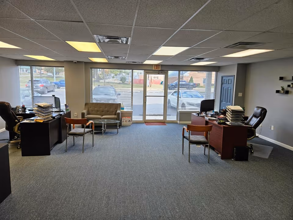 Office reception area with desks, chairs, stacks of papers, and large front windows.