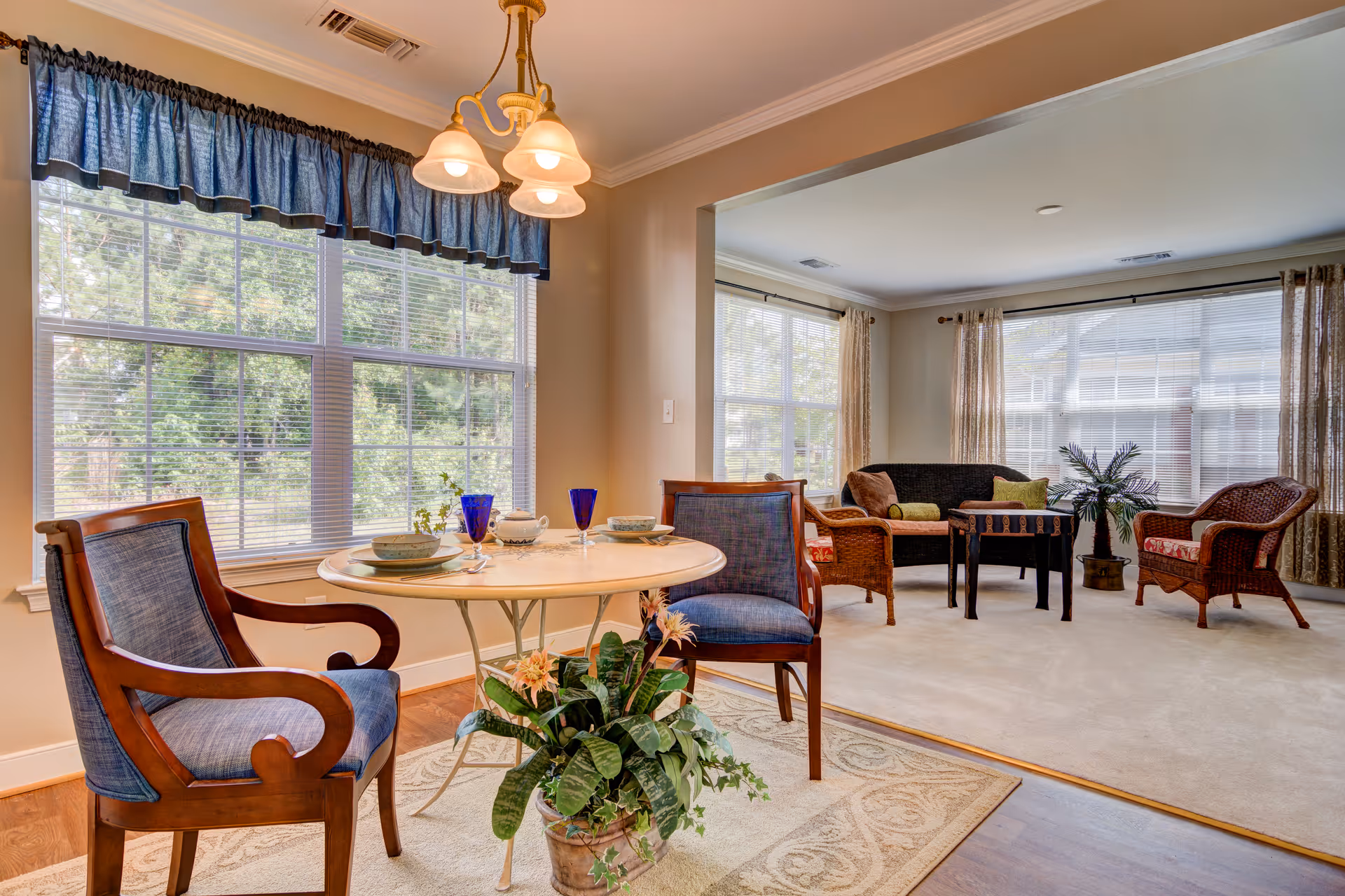 A bright and cozy interior space featuring a small round dining table set for two with blue cushioned wooden chairs and blue glassware. The dining area is adjacent to a living room with wicker furniture including a loveseat, two armchairs, a small black coffee table, and a potted plant. Large windows with blinds and curtains allow natural light to fill the room. A blue valance hangs above the dining area window, and a three-light chandelier is suspended above the table.