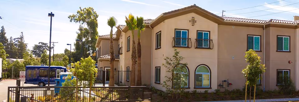 Front exterior of a two-story beige stucco memory care building with small balconies, palm trees, a fenced planter area, and a blue community shuttle parked nearby.