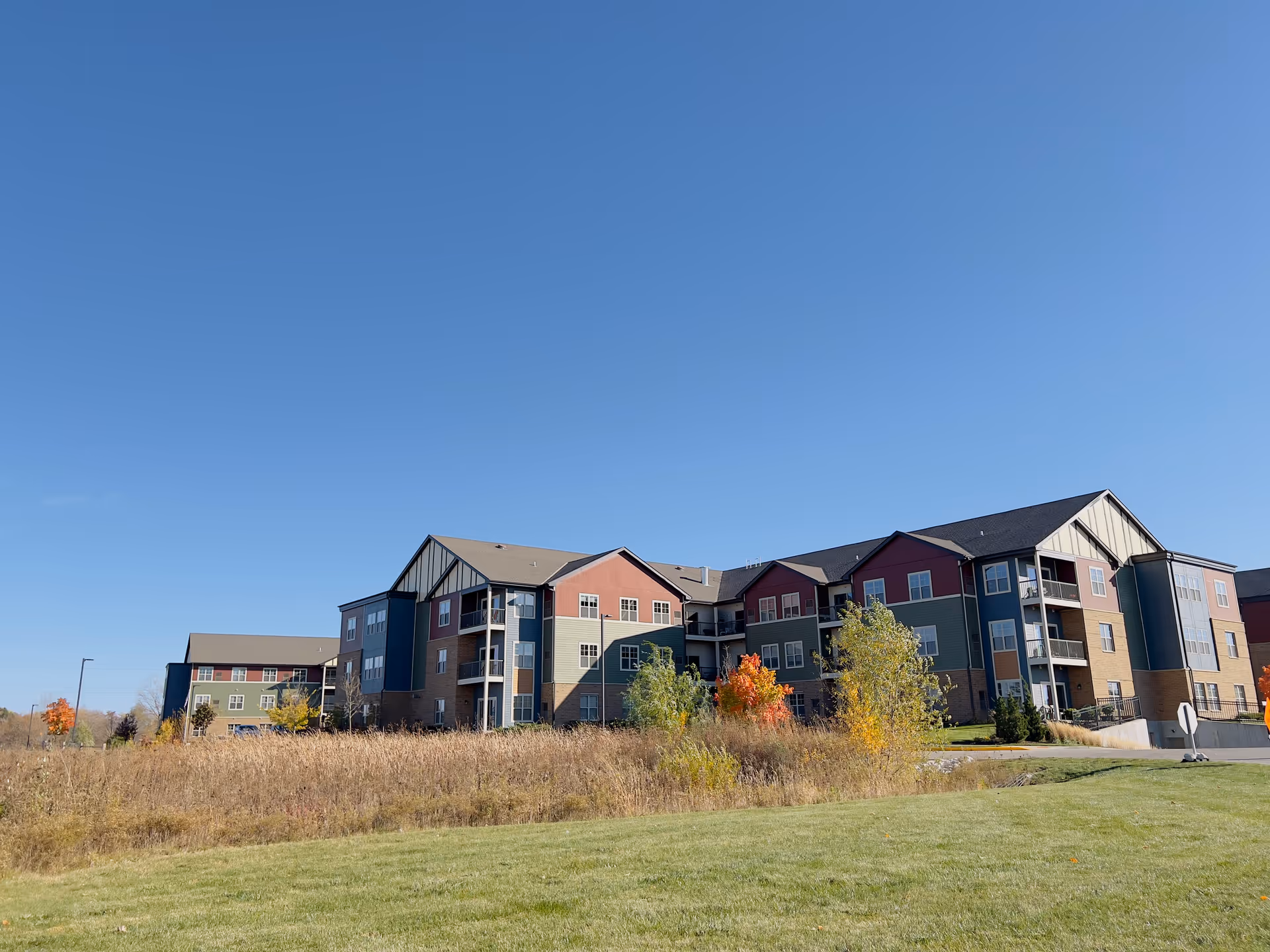 Exterior view of a multi-story residential building with a mix of red, green, and beige siding under a clear blue sky. The building is surrounded by grass, shrubs, and small trees with autumn foliage.