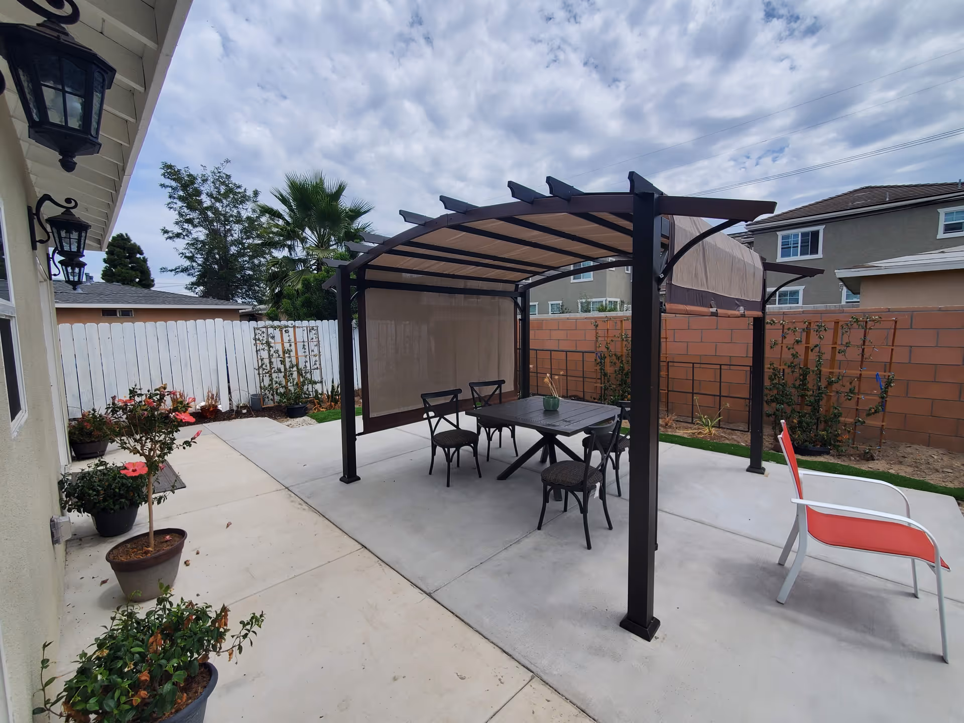 Backyard patio with a covered pergola over a dining table and chairs, potted plants, and surrounding fences.