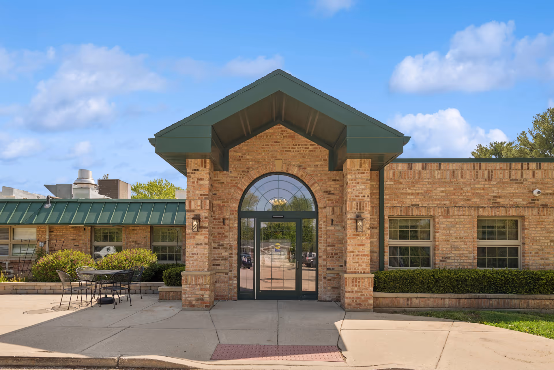 Brick entrance to senior living facility with arched glass doors and green gabled roof.