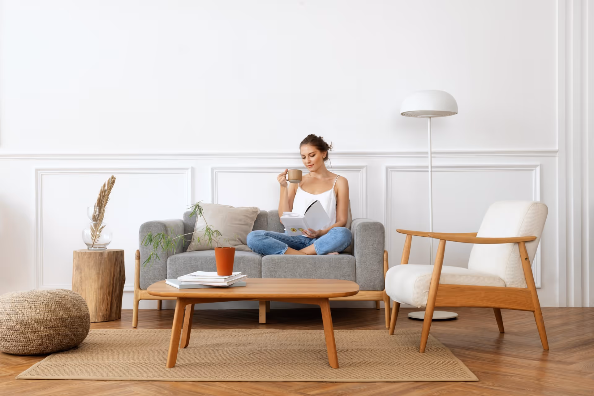 A woman sits on a gray sofa in a modern living room, reading and sipping coffee.