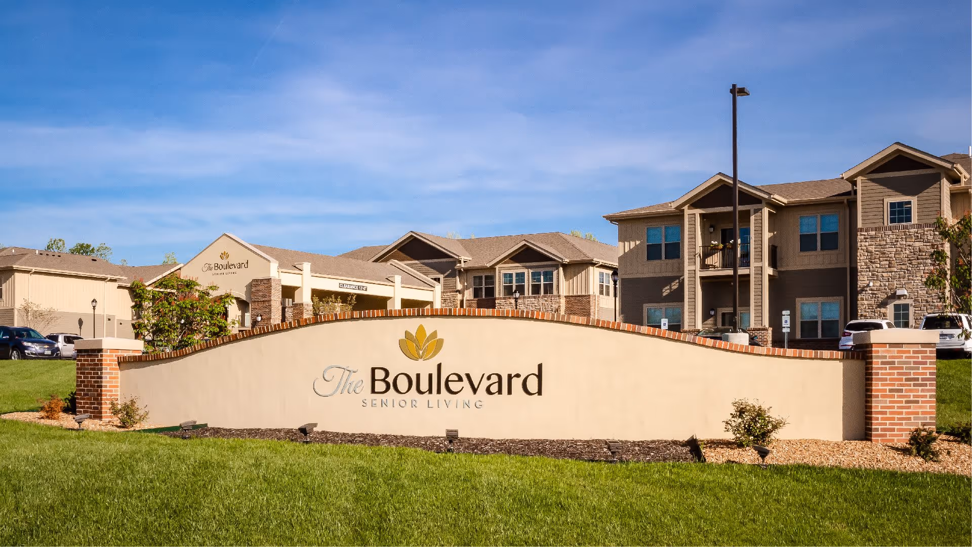 Exterior view of The Boulevard Senior Living facility with a large sign in the foreground displaying the facility's name. The building behind the sign is a multi-story residential structure with beige siding, stone accents, balconies, and several windows under a clear blue sky.
