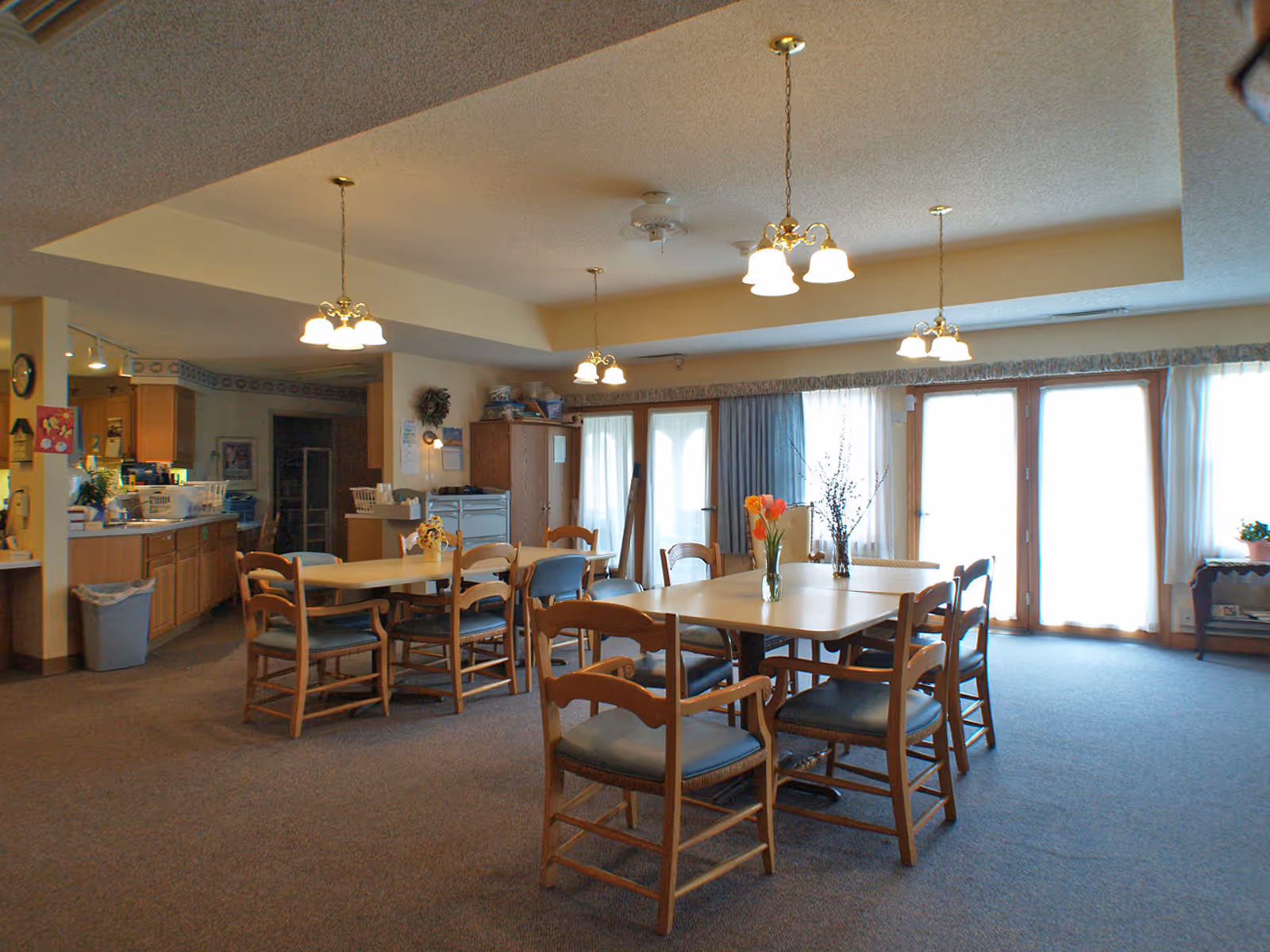 A dining area in a senior living facility with multiple wooden tables and chairs arranged on a carpeted floor. The room is well-lit with hanging light fixtures and natural light coming through large windows and glass doors covered with sheer curtains. There are flower vases on the tables and a kitchen area visible in the background.