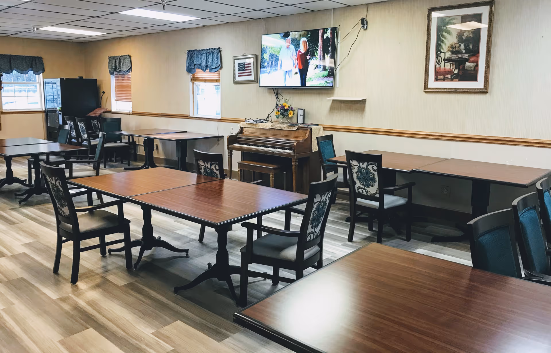 A dining room with several wooden tables and chairs arranged neatly. There is a piano against the wall beneath a mounted flat-screen TV displaying an outdoor scene. The room has light-colored walls with framed artwork and windows with blue valances allowing natural light to enter.