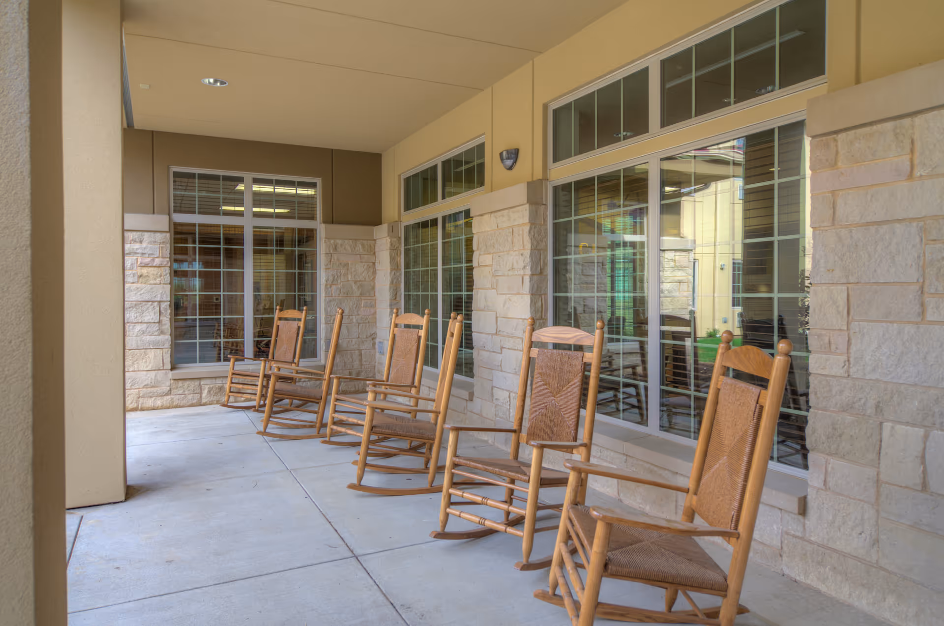 Row of wooden rocking chairs on a covered outdoor patio beside stone-walled building windows.