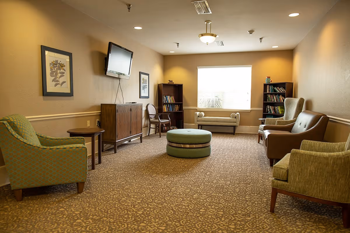 A cozy living room area with patterned carpet, several armchairs, a round ottoman in the center, two bookshelves filled with books, a wall-mounted flat screen TV, framed botanical artwork on the walls, and a window with blinds letting in natural light.