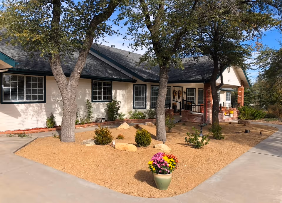 Senior living facility exterior with beige siding, trees, and colorful potted flowers.