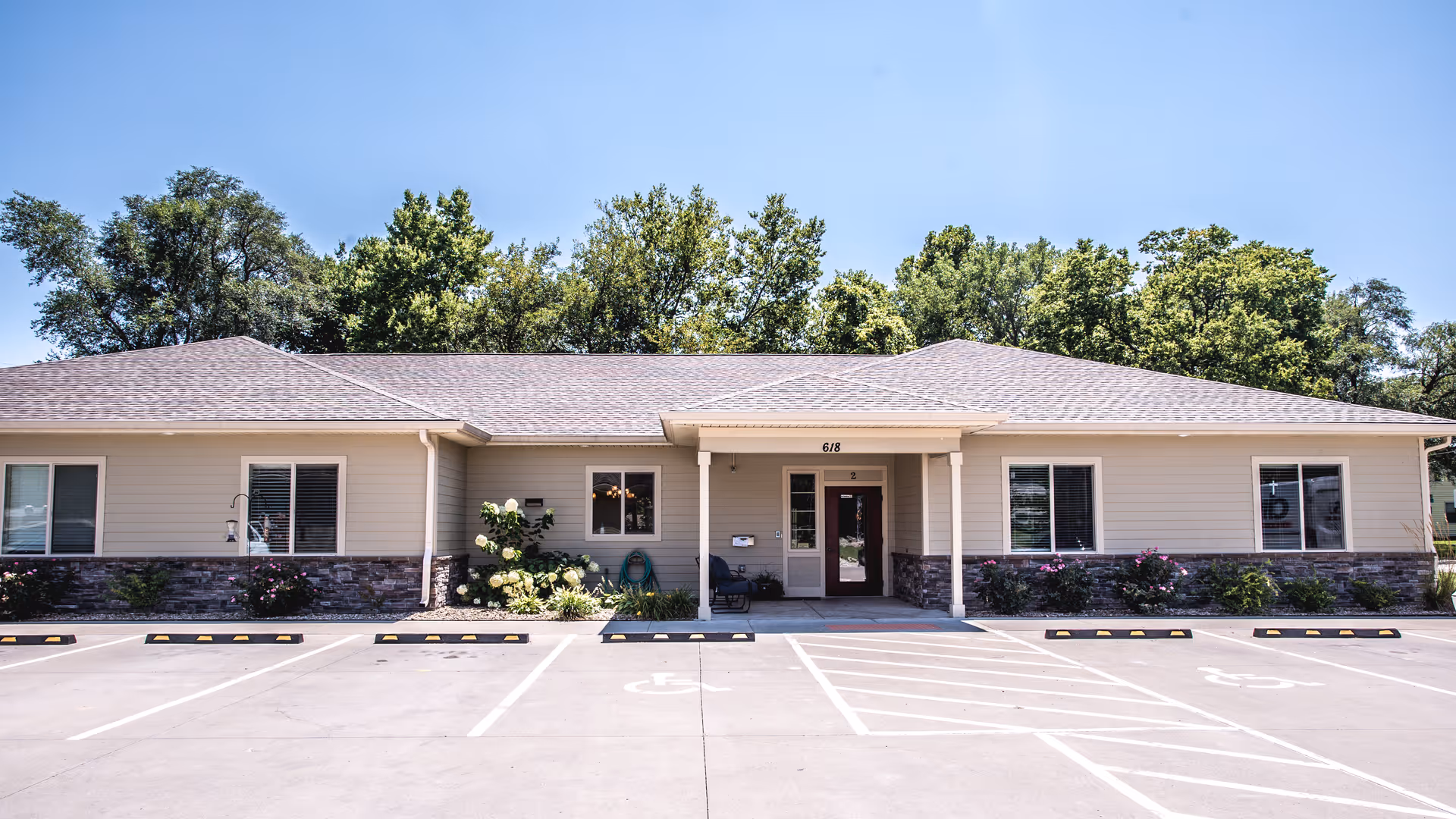 Single-story beige building front with a covered main entrance, landscaped flower beds, and empty parking spaces in front.