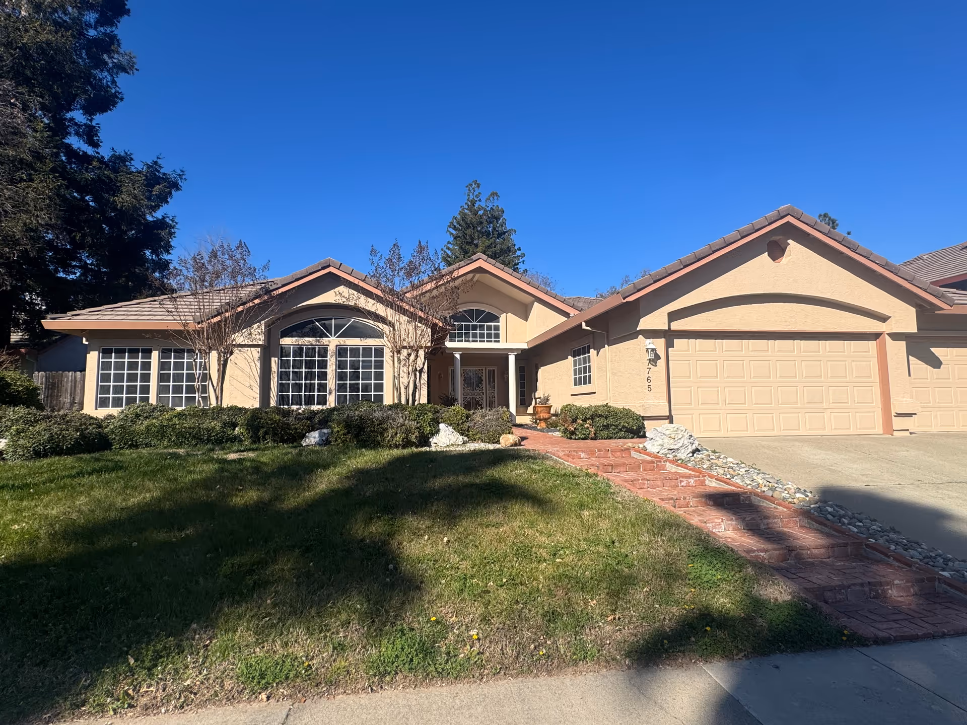 A beige, single-story residential building with a brick walkway, front yard, and garage.