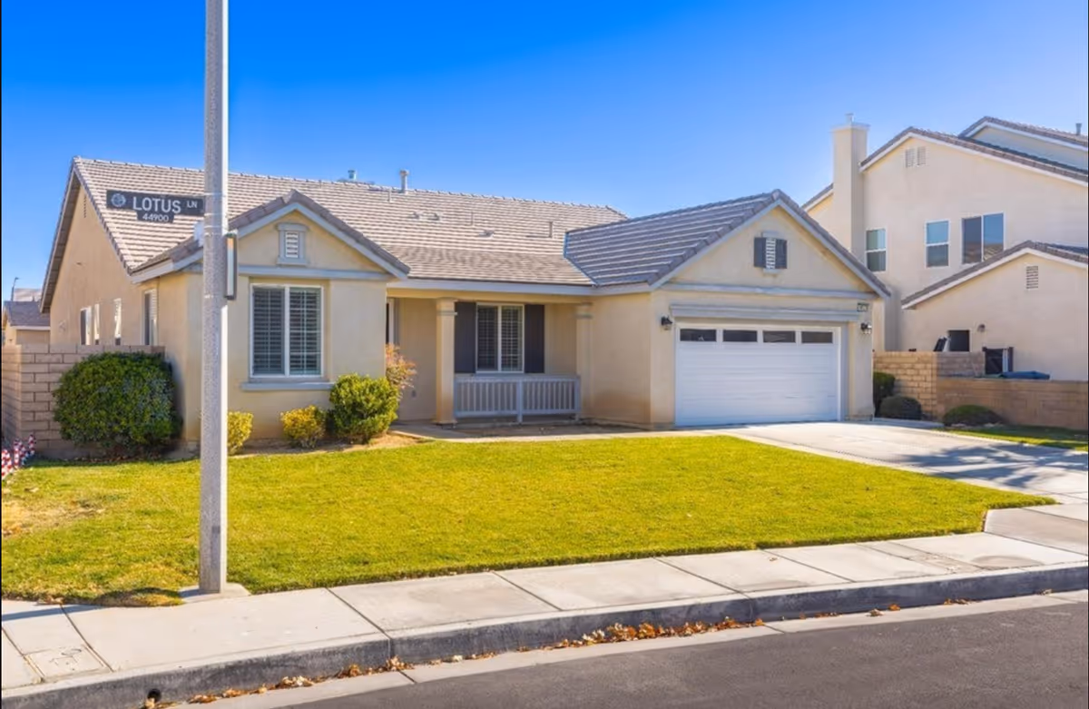 Single-story suburban house with a two-car garage, front porch and a green lawn under a clear blue sky.