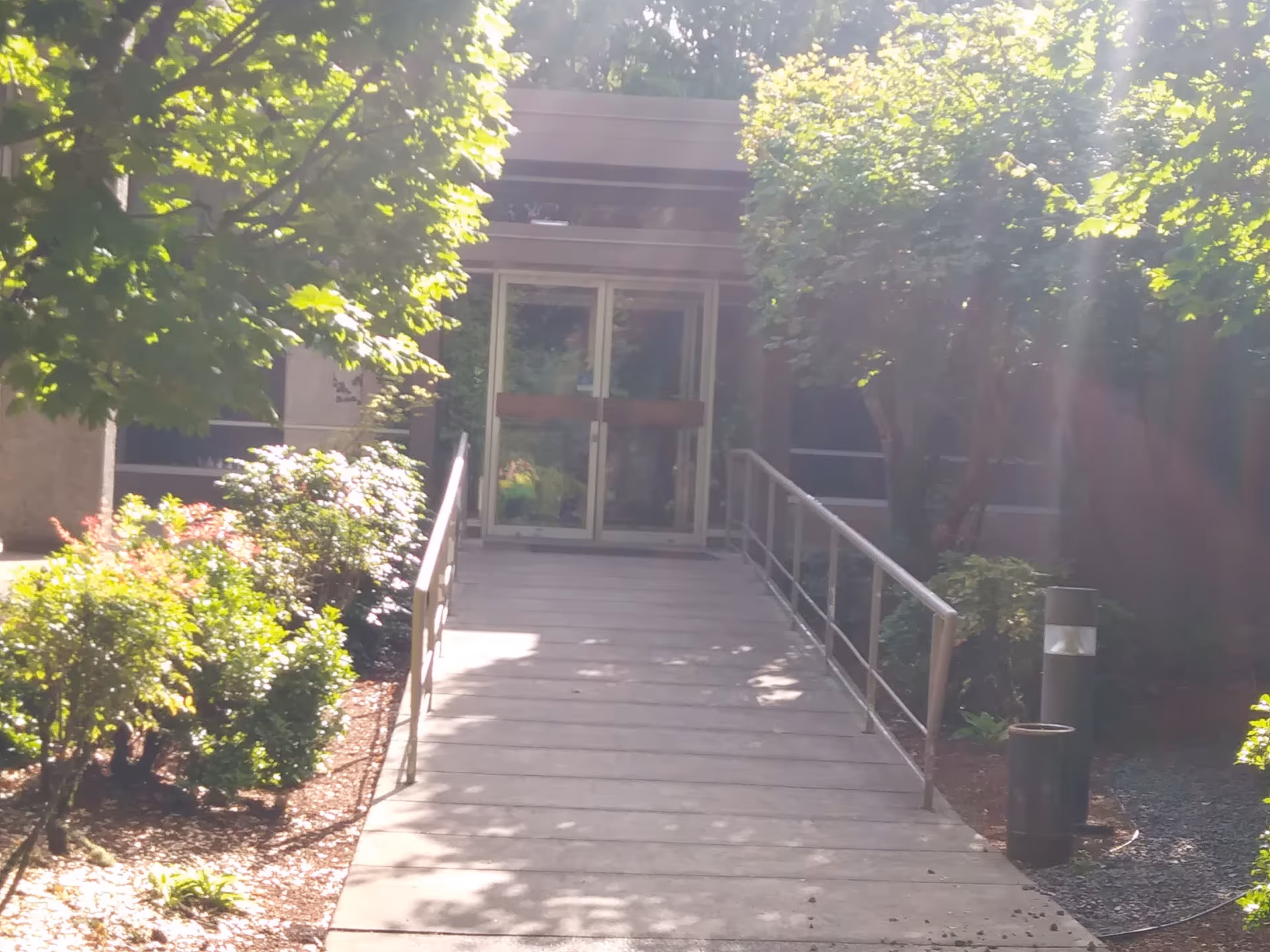 Front entrance with glass doors and a ramp, surrounded by trees and shrubs.