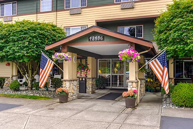 Entrance of a senior living facility with a covered porch supported by stone pillars, two American flags on either side, hanging flower baskets, potted plants, and a glass door under the address number 12806.