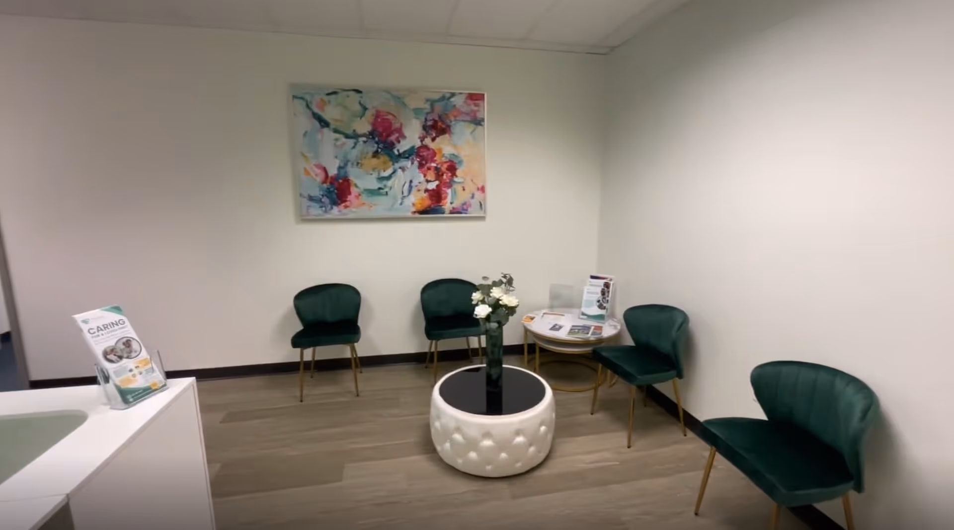 Small interior waiting area with green chairs around a round tufted white ottoman and abstract art.