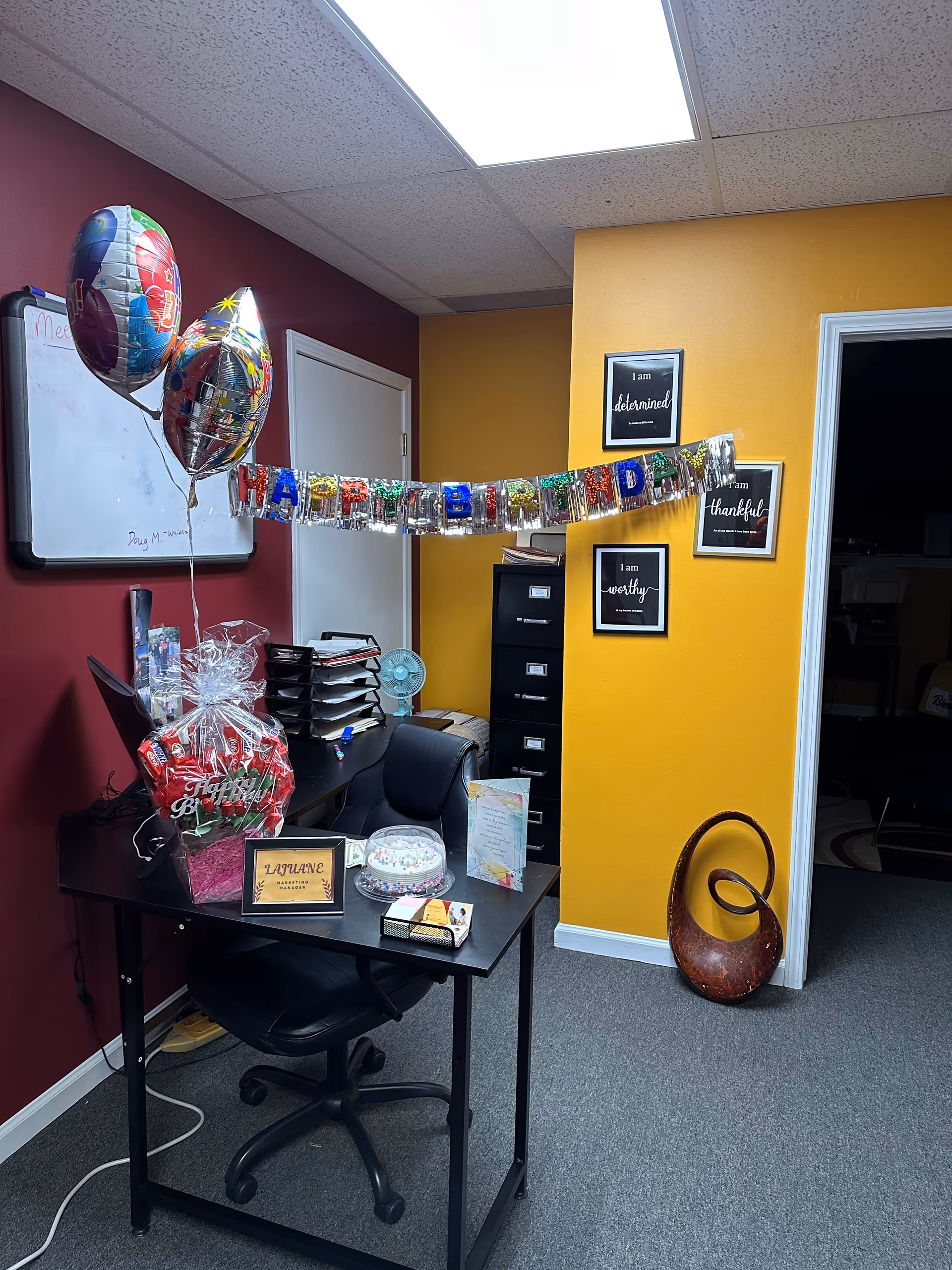 Office desk with birthday decorations, balloons, cake, and filing cabinet in a colorful room.