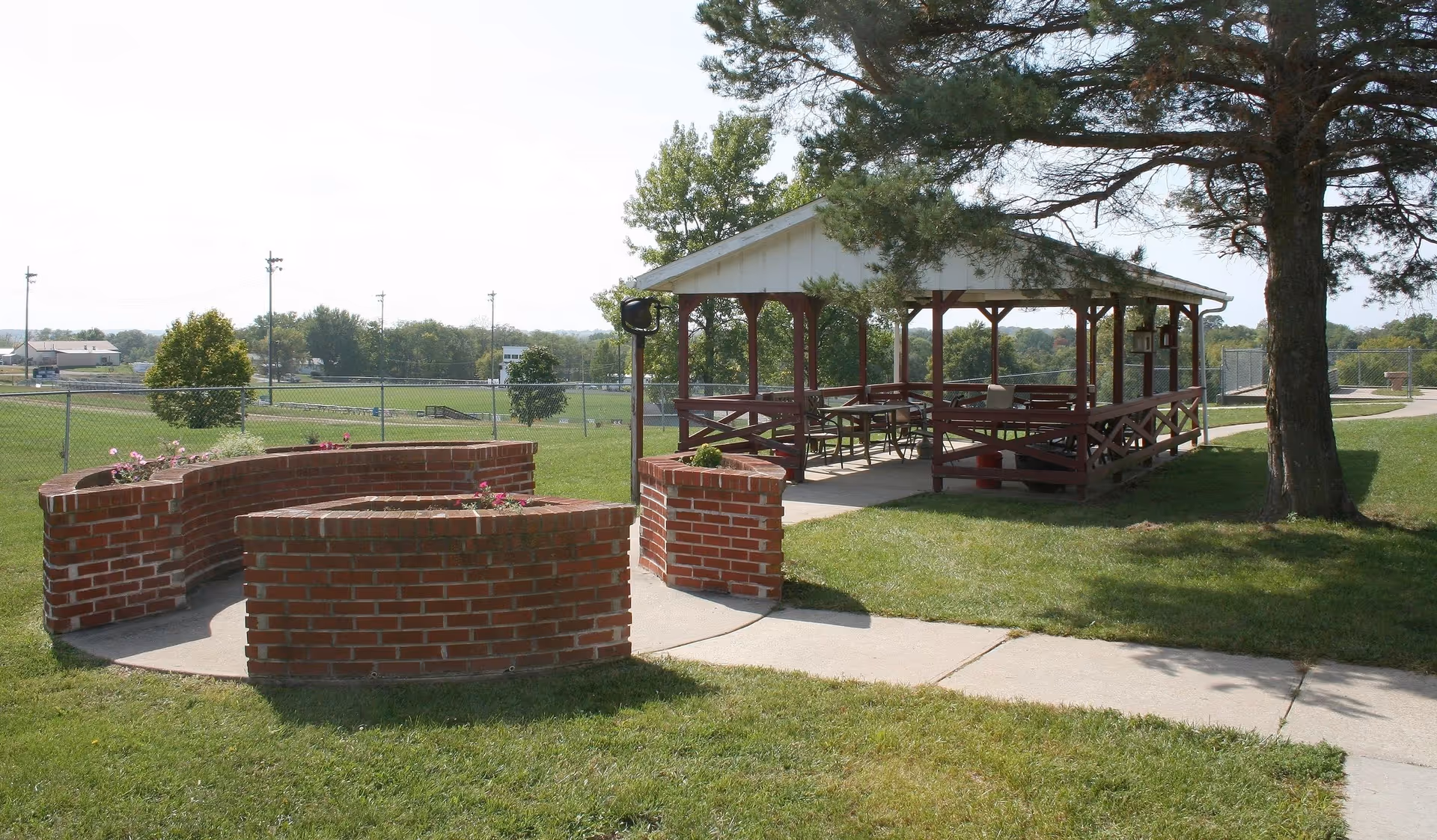 A grassy outdoor area with a covered wooden pavilion, curved brick planters, and a sidewalk under a large tree.