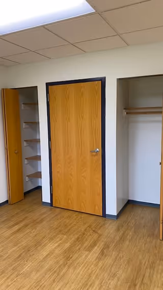Empty room with wood flooring, a central wooden door flanked by open closets and shelving under a drop ceiling.
