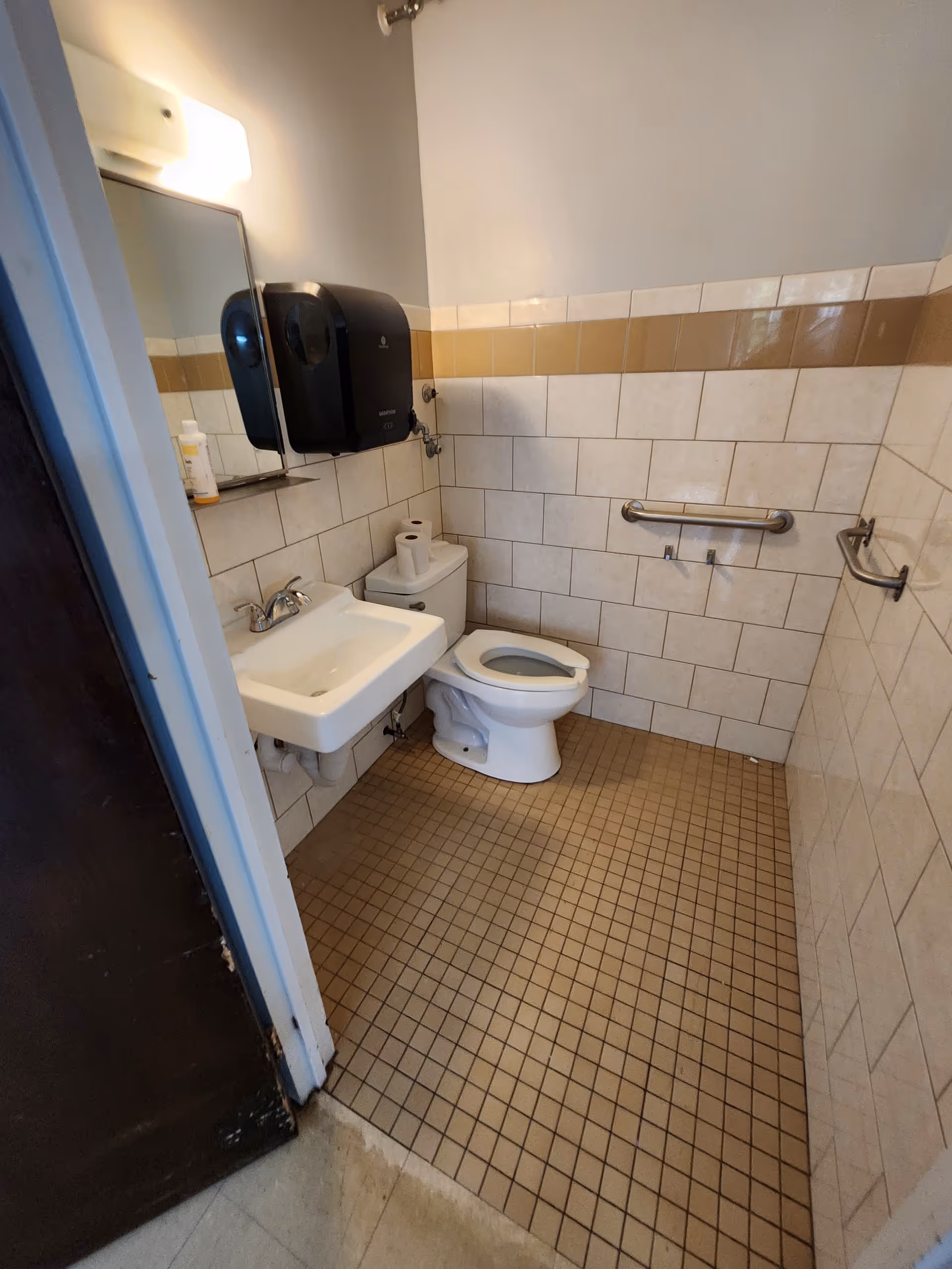 Small tiled bathroom with sink, toilet, grab bars, and wall-mounted soap dispenser.