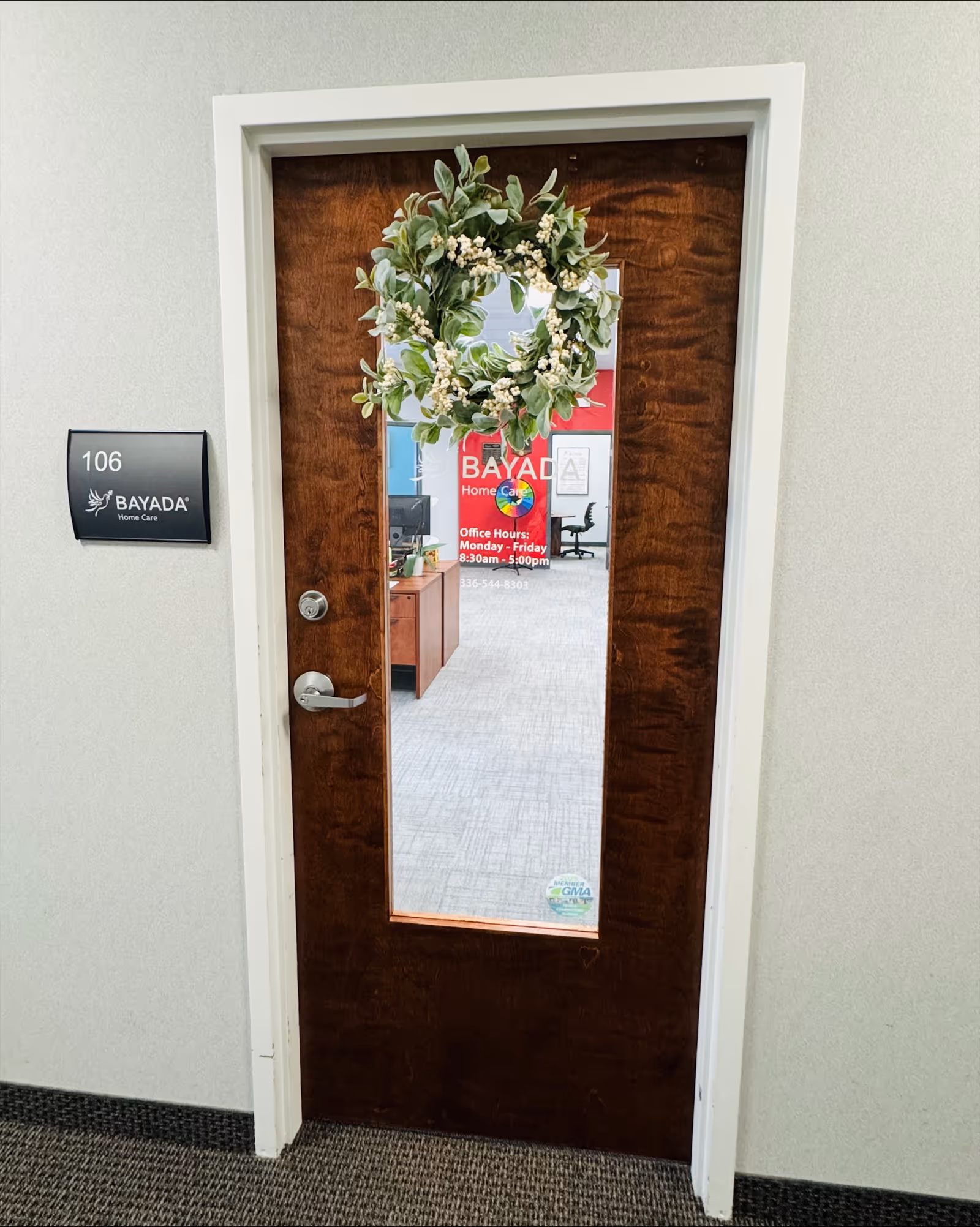 Wooden interior door with wreath, BAYADA sign, 106 plaque, and office visible through glass panel.