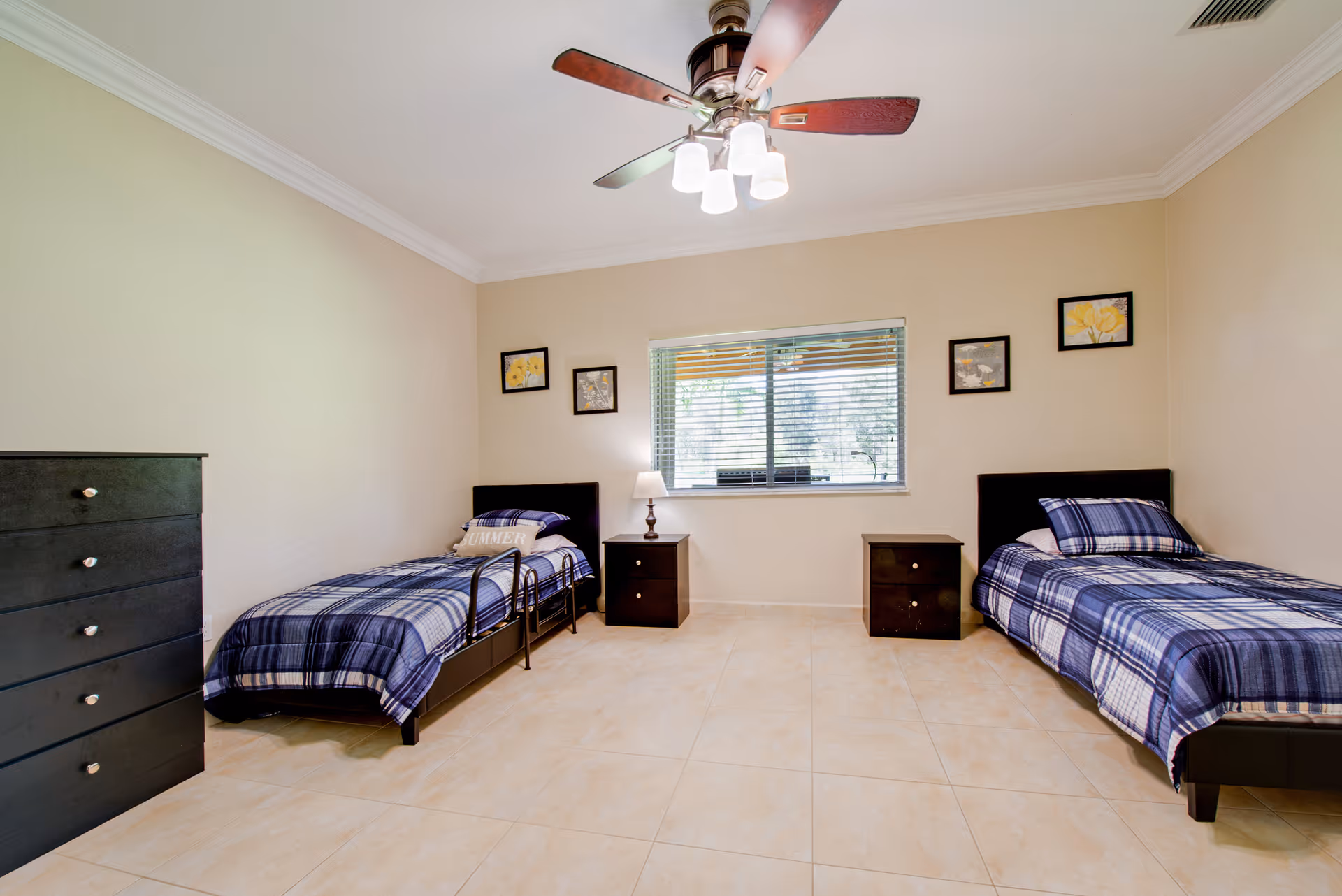 A clean and simple bedroom with two single beds on opposite sides of the room, each with blue plaid bedding. Between the beds are two dark wooden nightstands, one with a lamp. A window with blinds is centered on the back wall, and four small framed pictures hang on the walls. A dark wooden dresser is on the left side of the room. The floor is tiled, and a ceiling fan with lights hangs above.