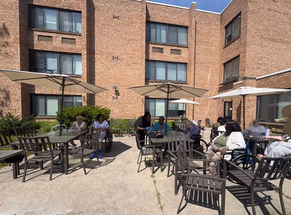 Group of seniors seated at outdoor tables under umbrellas beside a brick building, some in wheelchairs.